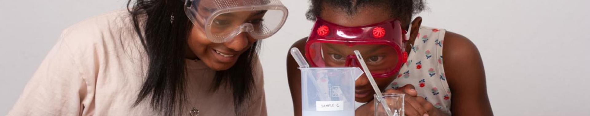 Two smiling girls in goggles observing a chemical reaction in a beaker.